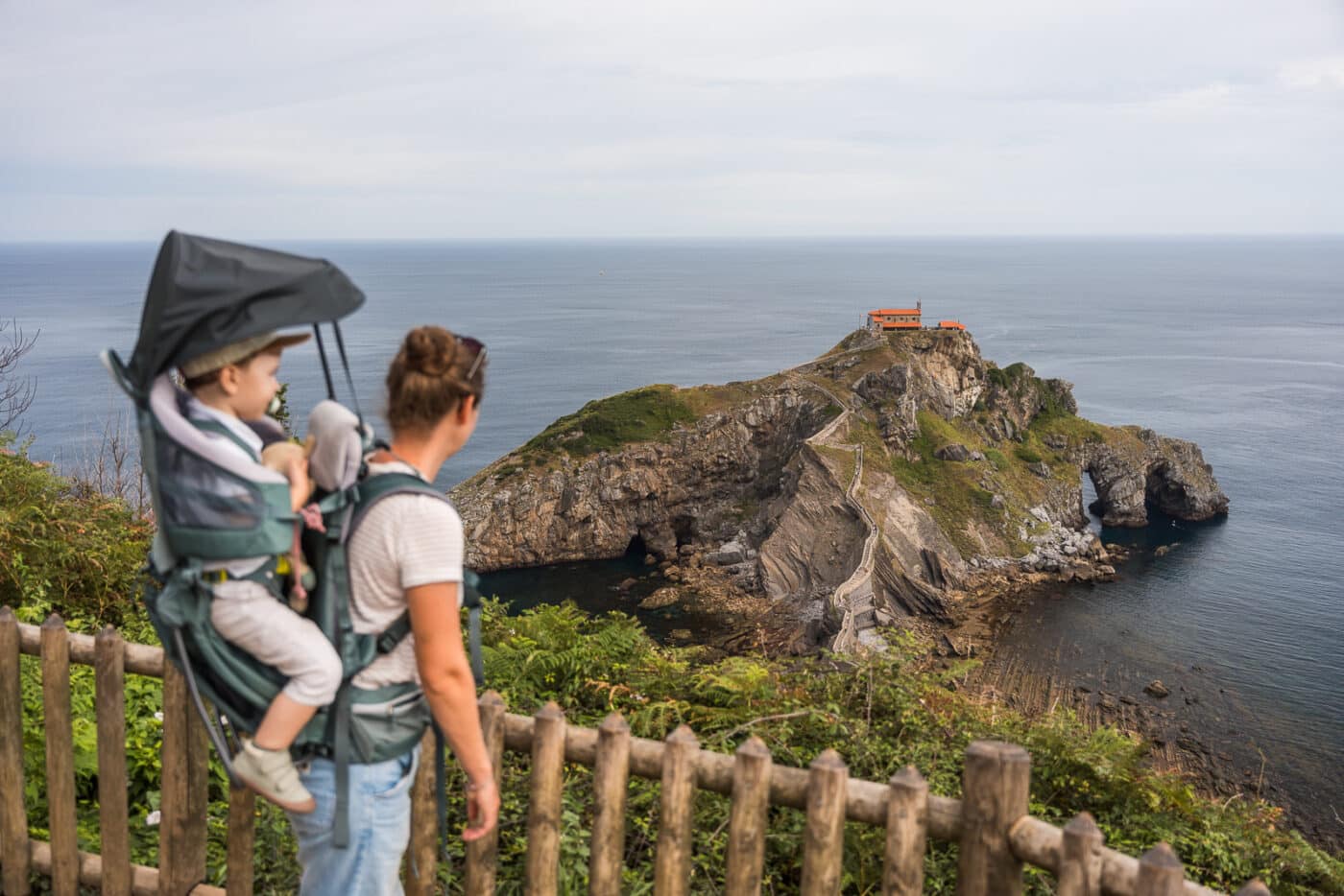 Adulte avec un enfant en porte-bébé face à San Juan de Gaztelugatxe, avec l’escalier et l’ermitage en arrière-plan.