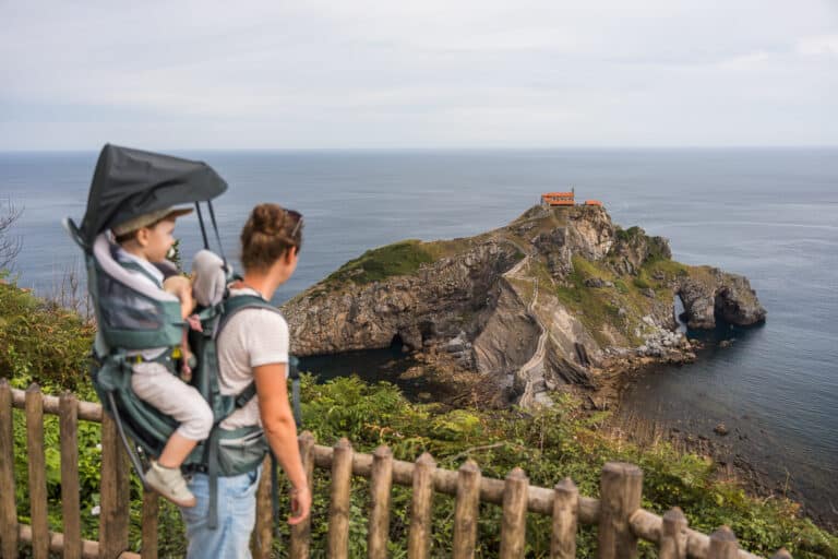 Adulte avec un enfant en porte-bébé face à San Juan de Gaztelugatxe, avec l’escalier et l’ermitage en arrière-plan.